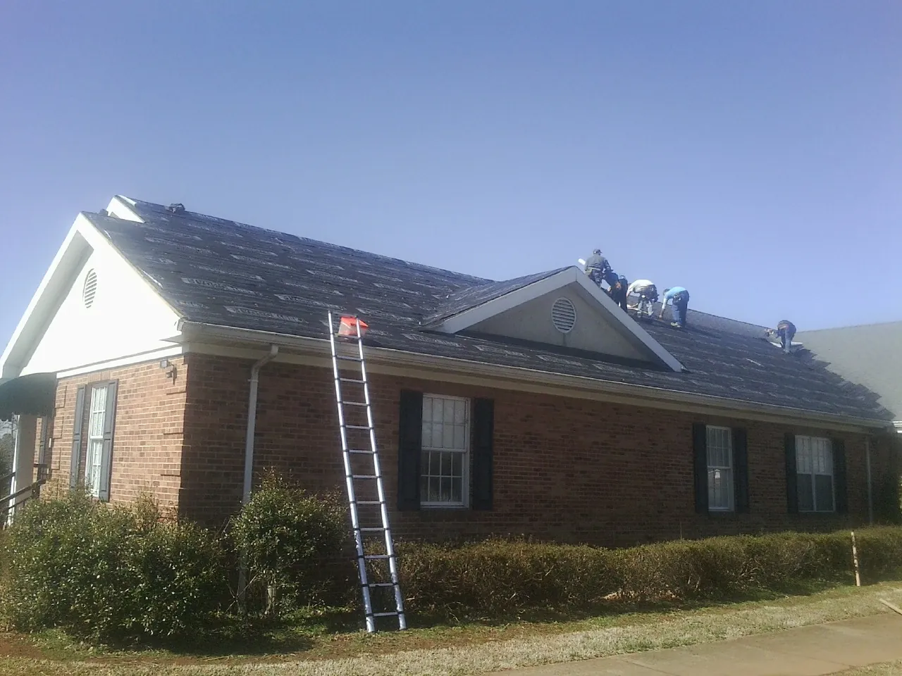 Crew tearing shingles off a brick ranch roof with a ladder set — Emergency Roof Repair work by our crew in Minetto
