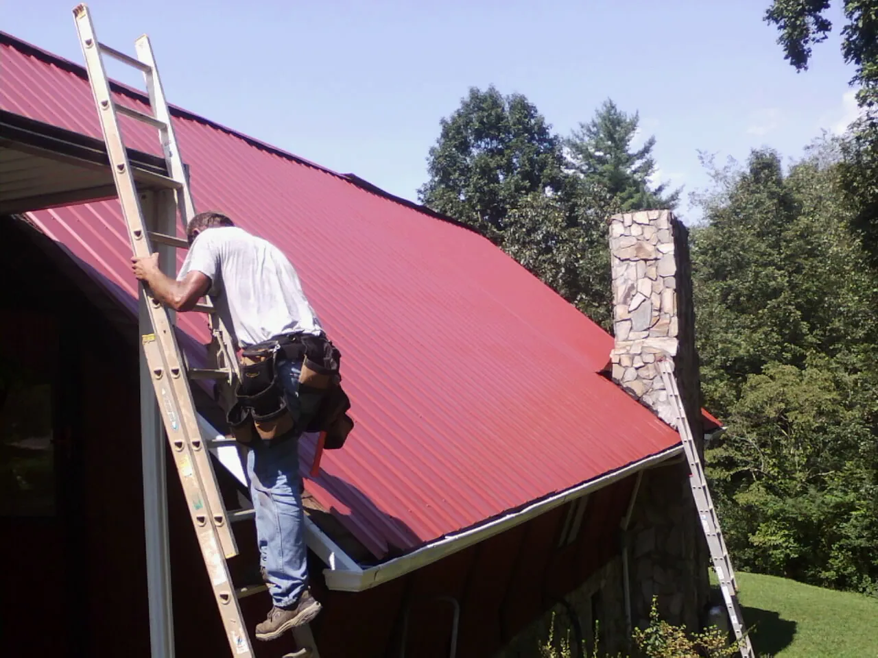 Roofer on a ladder beside a red metal roof and stone chimney — Soffit & Fascia Repair by Roofing Brothers in Minetto